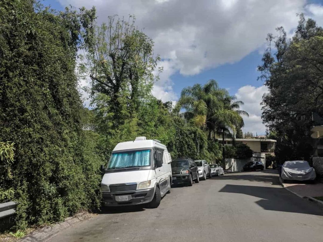 Van with sunshades in the front window parked next to a tall hedge in a residential neighborhood