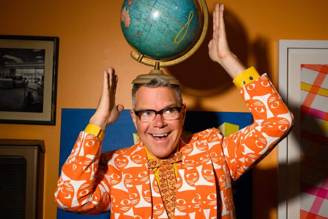 Charles Phoenix, in a bright orange cat-print suit, poses with a vintage globe on his head.