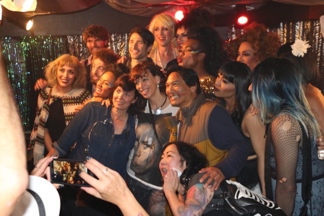 A crew of drag queens pose together on stage