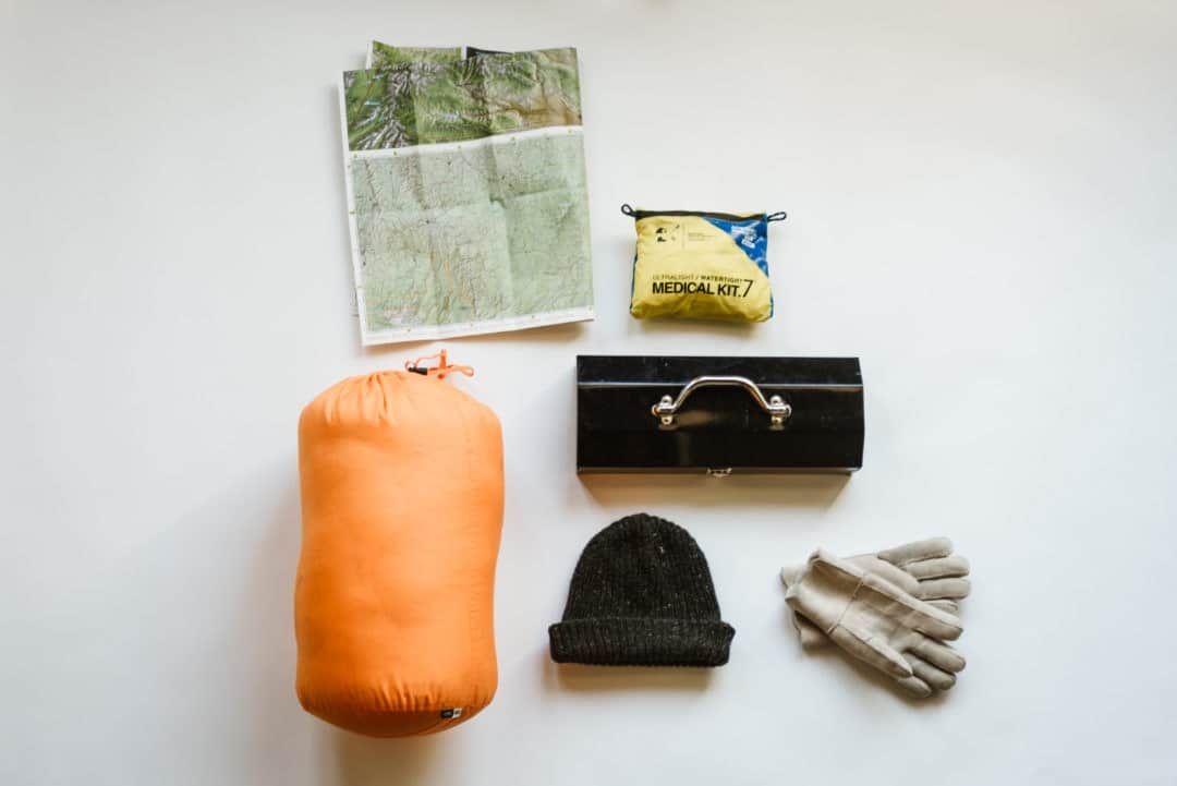 Camping gear laid out including a sleeping bag, map, gloves, medical kit, hat, and toolbox