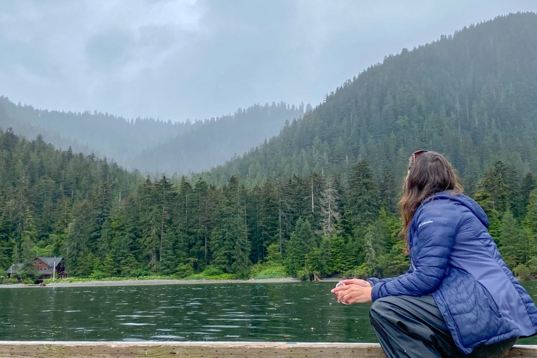 A woman with her back toward the camera looking out over a landscape of water and tree-covered mountains