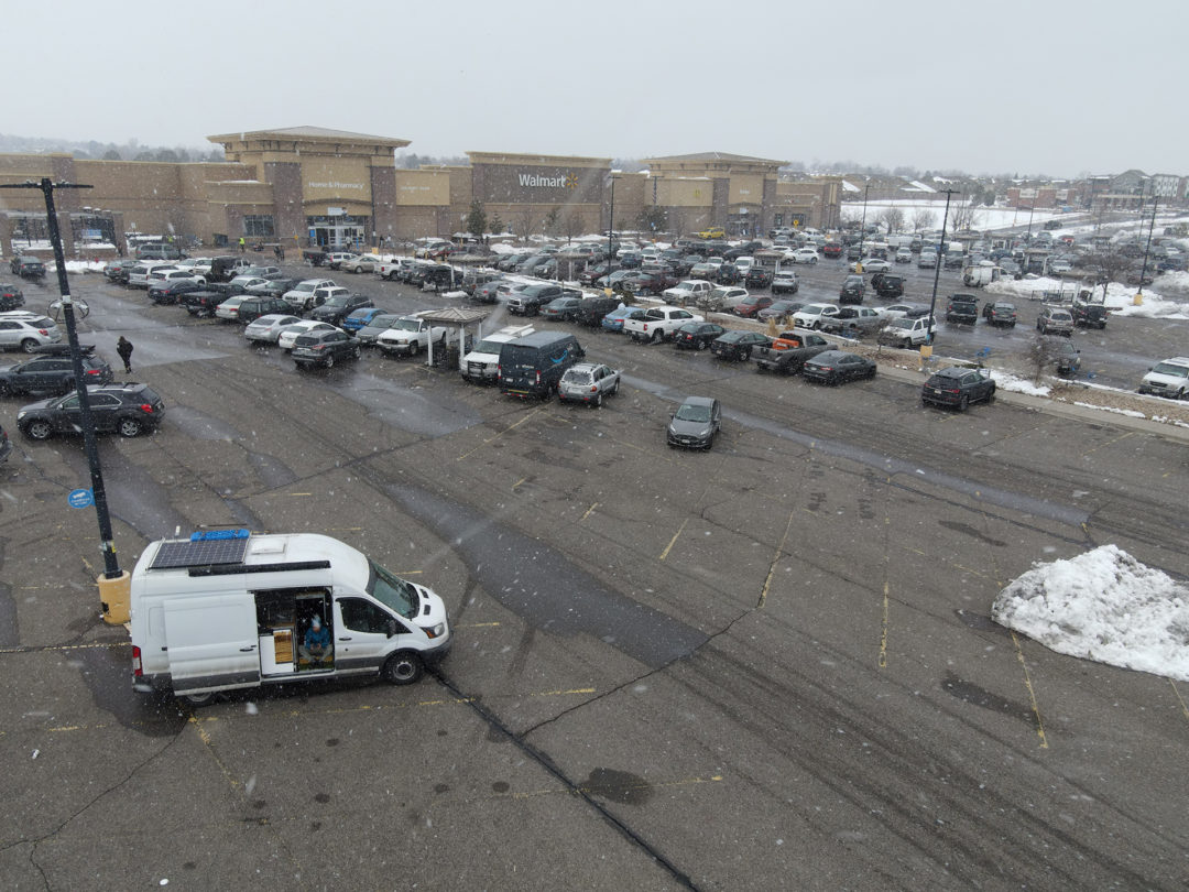 A white van is parked in a full Walmart parking lot as it snows
