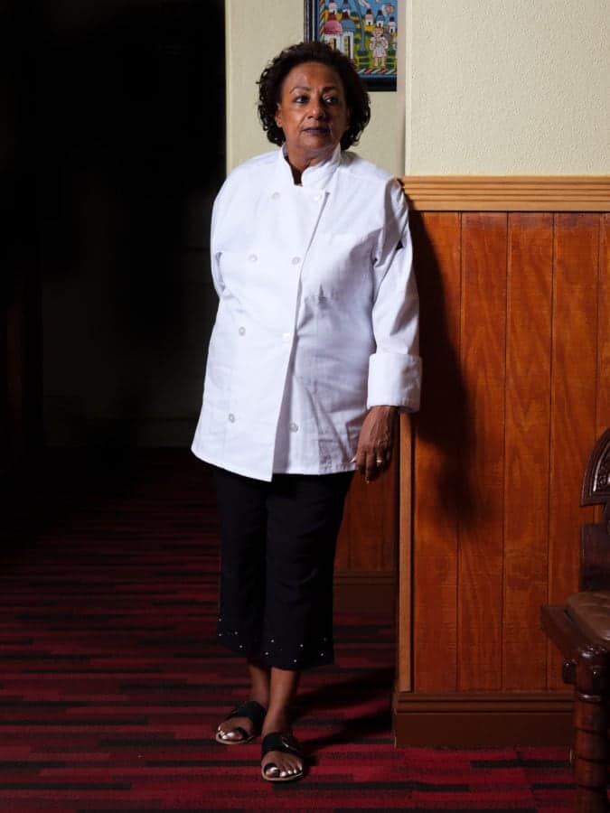 Women in white chef's jacket leans against wood-paneled wall and looks off to the right of the camera