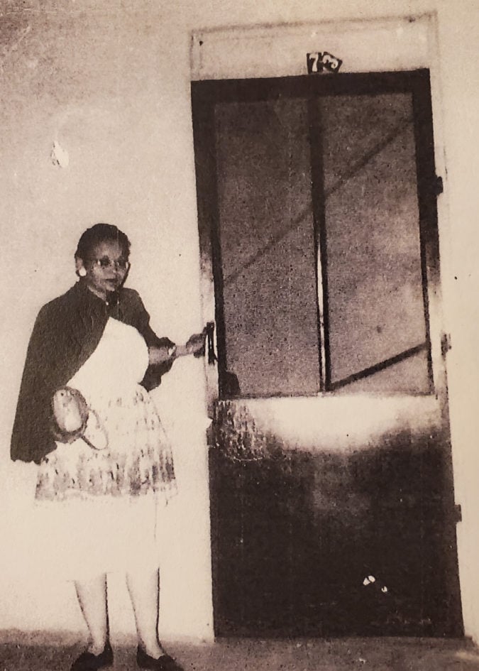 a black woman wears a white dress, glasses and a shawl and stands in front of a door