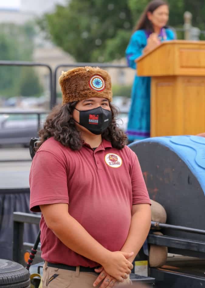 A man in a fur hat stands guard over the totem pole