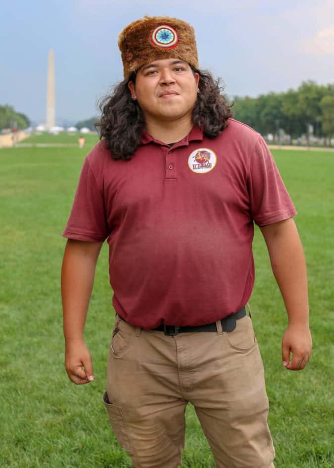 A man in a fur hat and maroon shirt stands on the national mall with the washington monument in the background