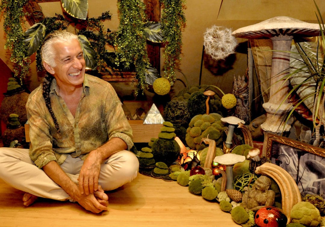 A smiling man sits cross-legged in a room surrounded by mossy plants