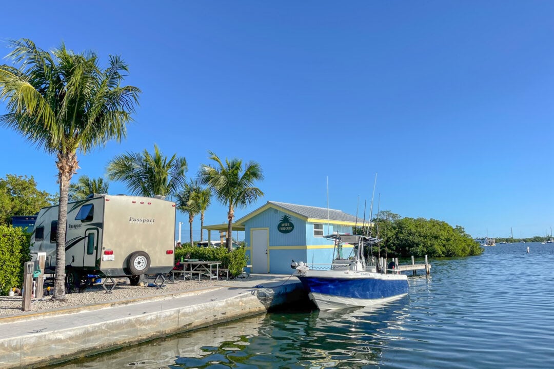 an RV is parked next to the water with a boat nearby, palm trees, and blue skies