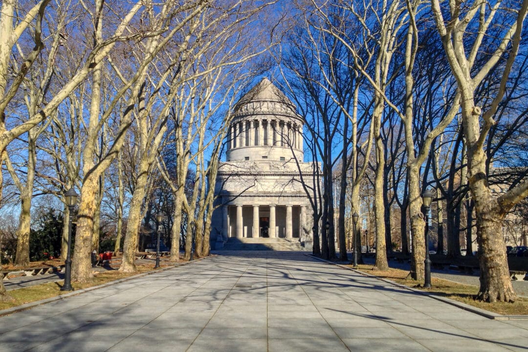 a large granite mausoleum set against a blue sky with a row of bare trees and wide open plaza