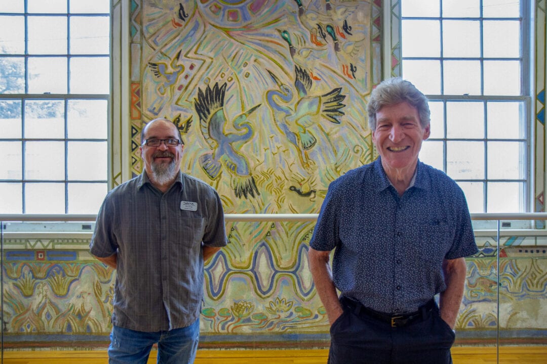two men pose smiling in front of a brightly colored mural