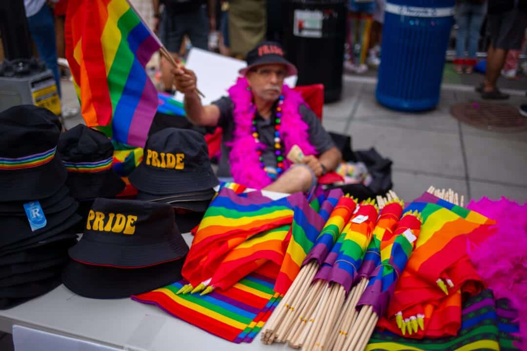 a vendor sells pride merchandise and wears a pink feather boa while waving a pride flag