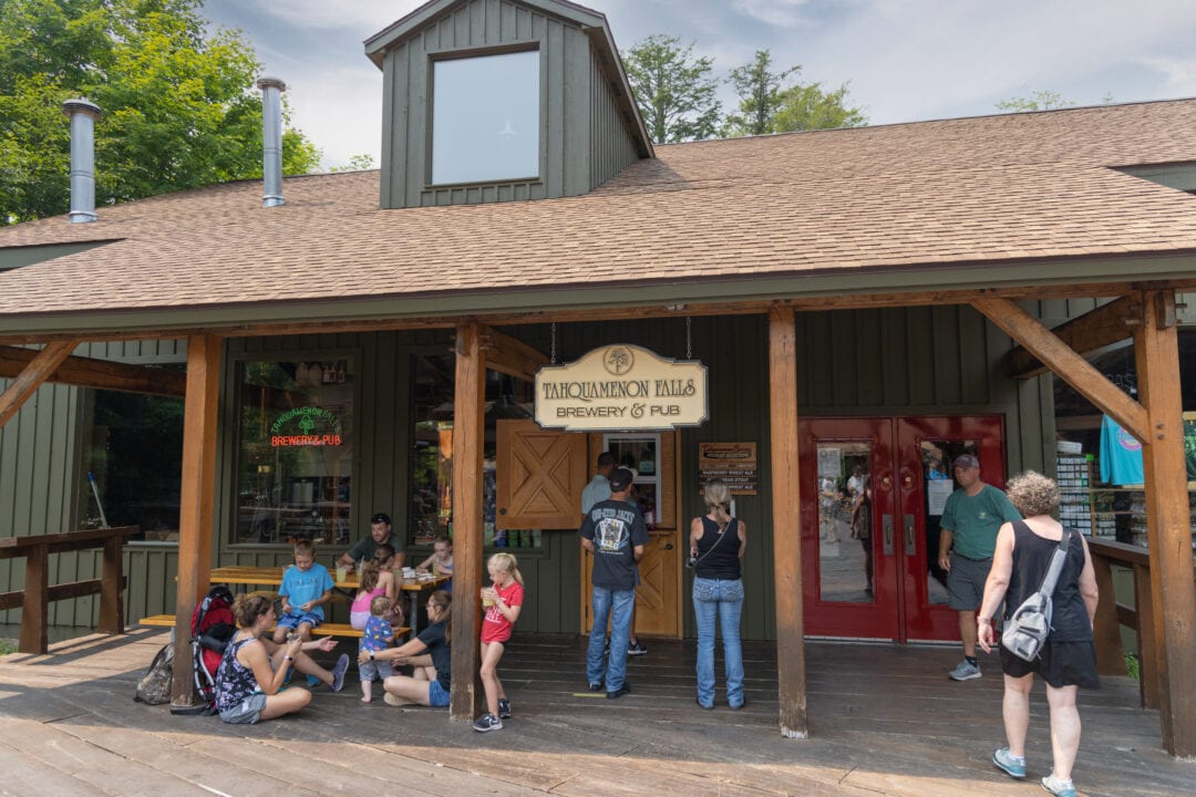 people wait outside of a brewery in a one-story wooden building painted green with a red door