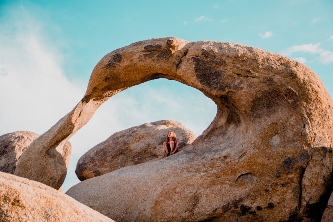 a child sits under a beige stone arch under a blue sky
