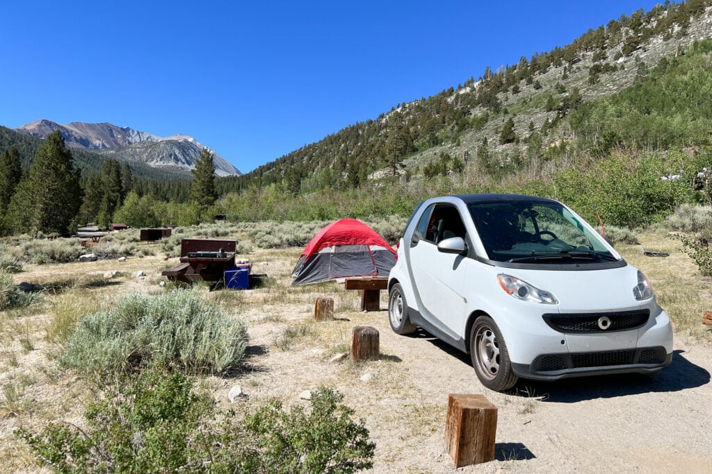 a white smart car parked at a campsite with a red tent popped behind it