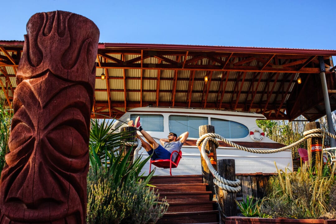 a person sits in a red chair next to a boat on land in front of a wooden tiki sculpture under a clear blue sky
