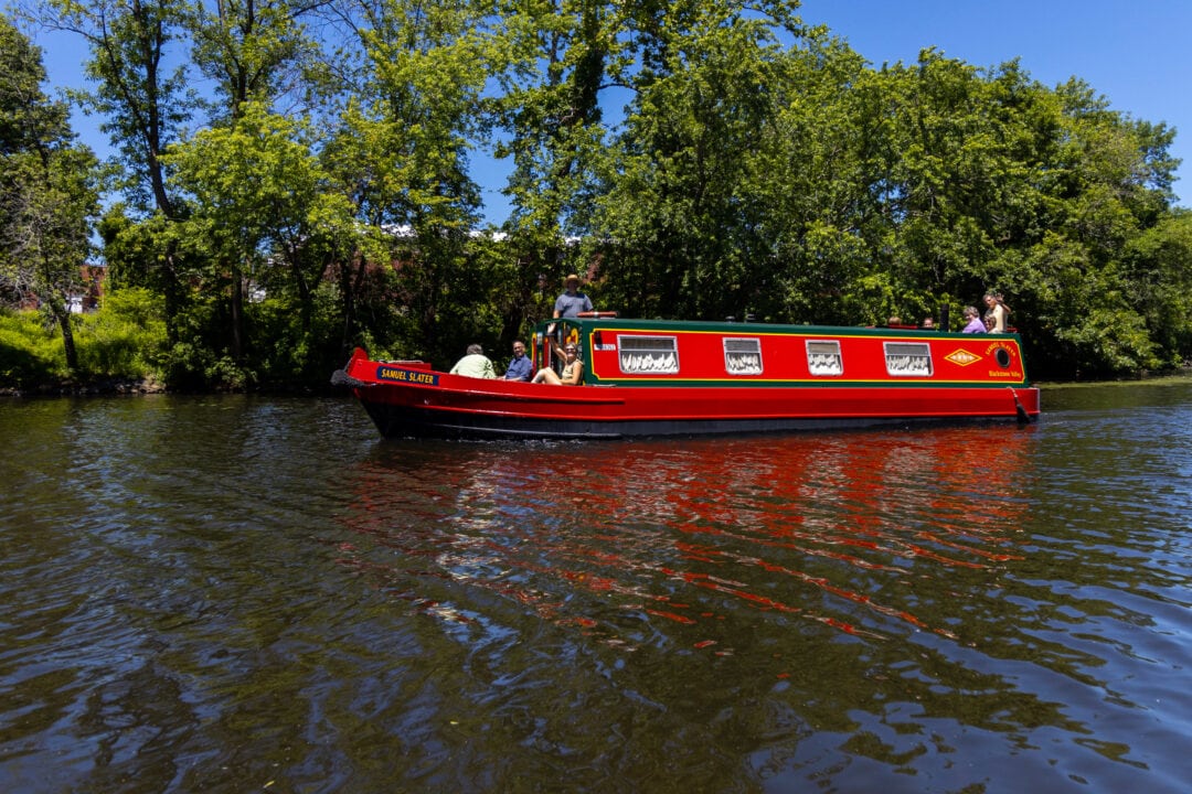 a small red boat floats on a river set in front of greenery and a clear blue sky