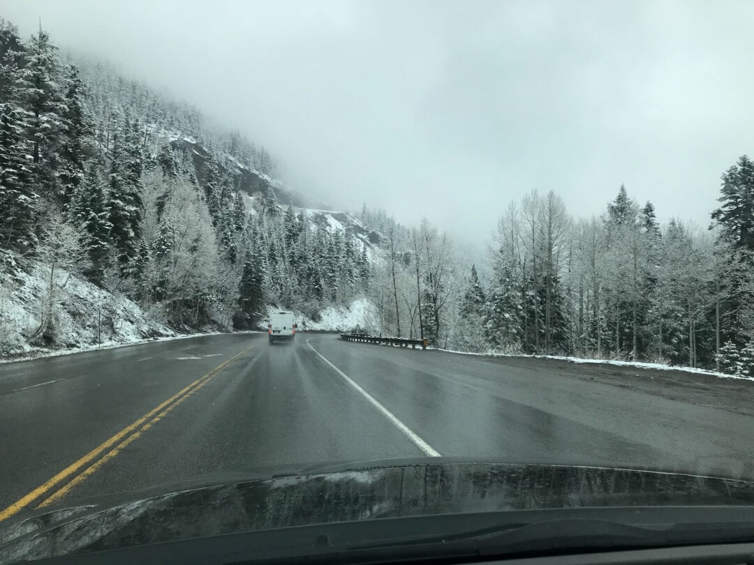 Wet road conditions and limited visibility cloud the views of snowy trees and mountains.