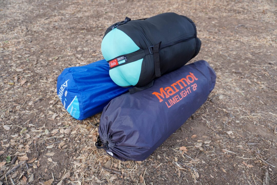 A tent, sleeping bag, and sleeping pad in their respective bags piled up on a dirt surface