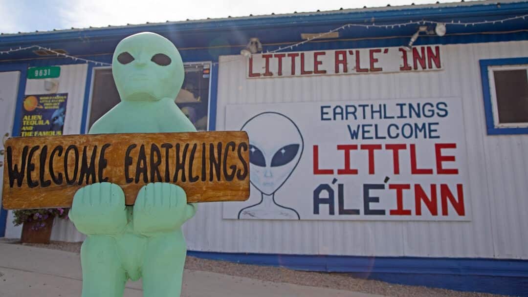 A green alien statue holds a sign that reads 'Welcome Earthlings' In front of a blue and white inn