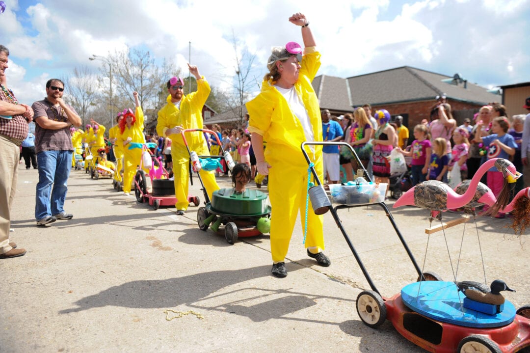 Partygoers dressed in matching yellow jumpsuits march in formation, each pushing a colorful lawn mower.