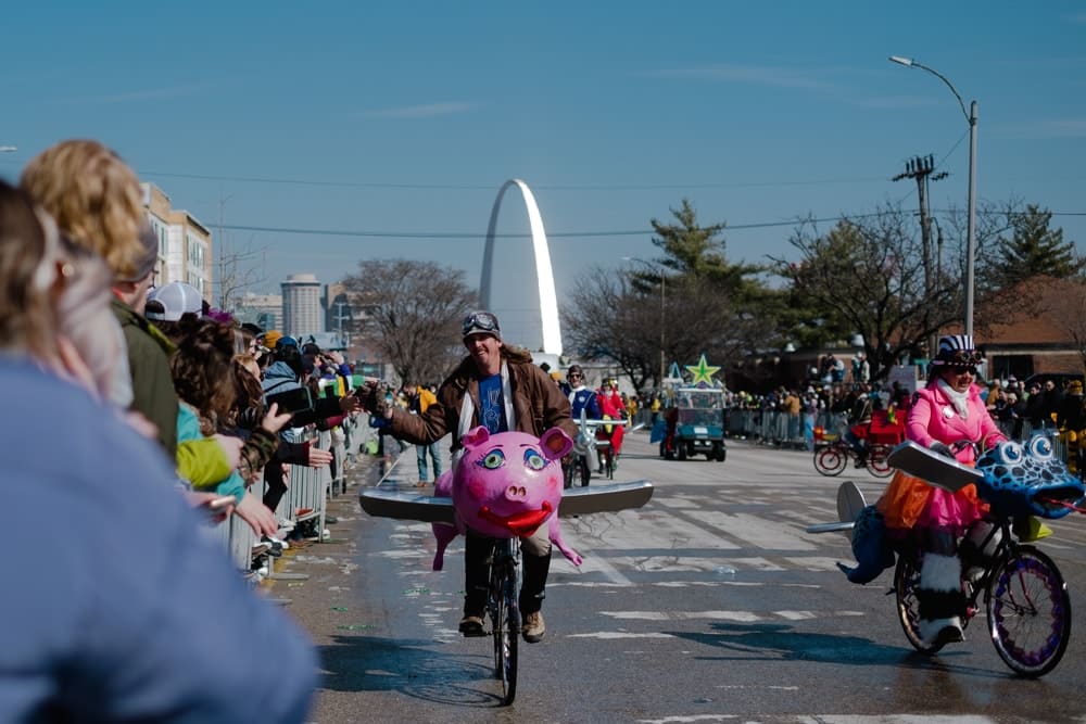 People riding bikes decorated as pigs and other animals glide down a roadway with St. Louis' Gateway Arch in the background.