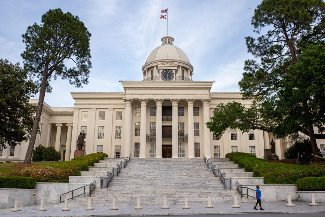 a three story white building with columns and a central dome flying two flags with a long granite staircase flanked by trees