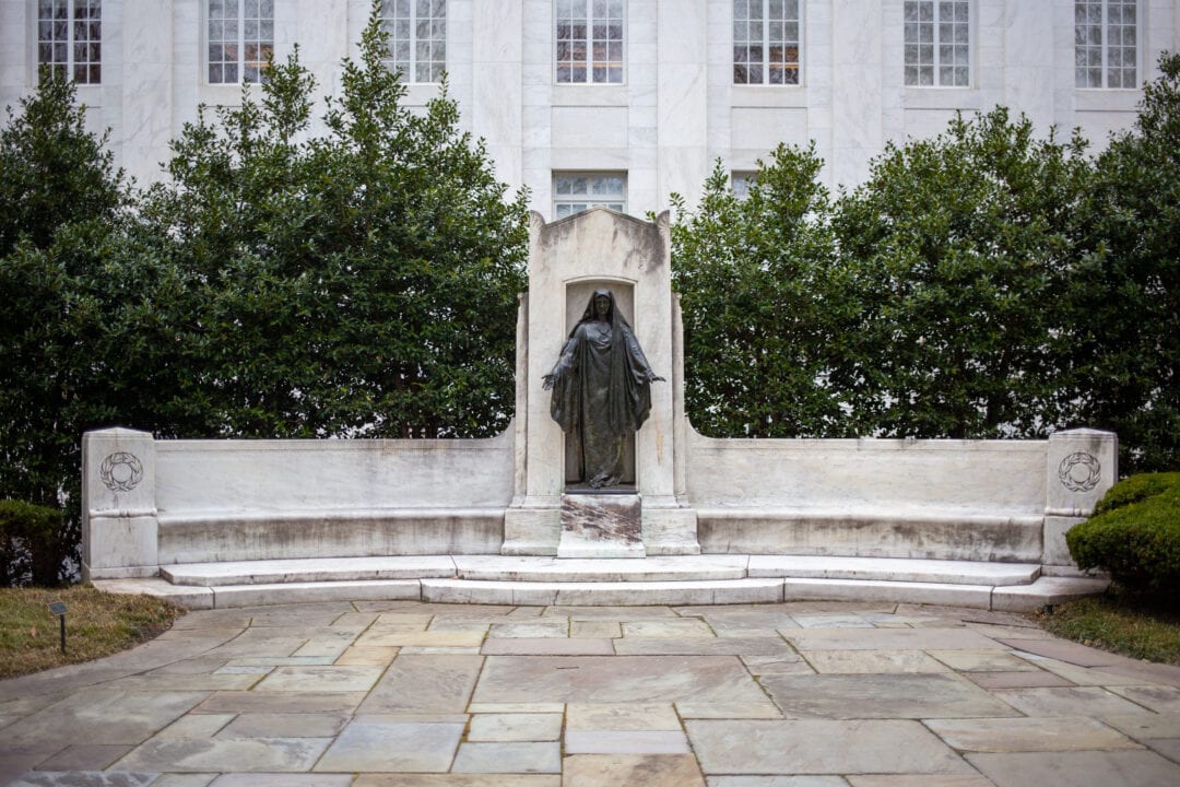 a bronze robed figure statue stands in the middle of a long white stone bench in the middle of a plaza
