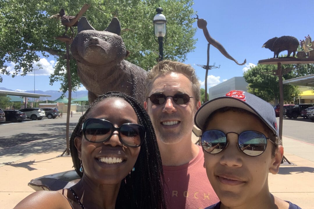 three people wearing sunglasses, including a black woman, white man and young child take a selfie in front of metal sculptures outside